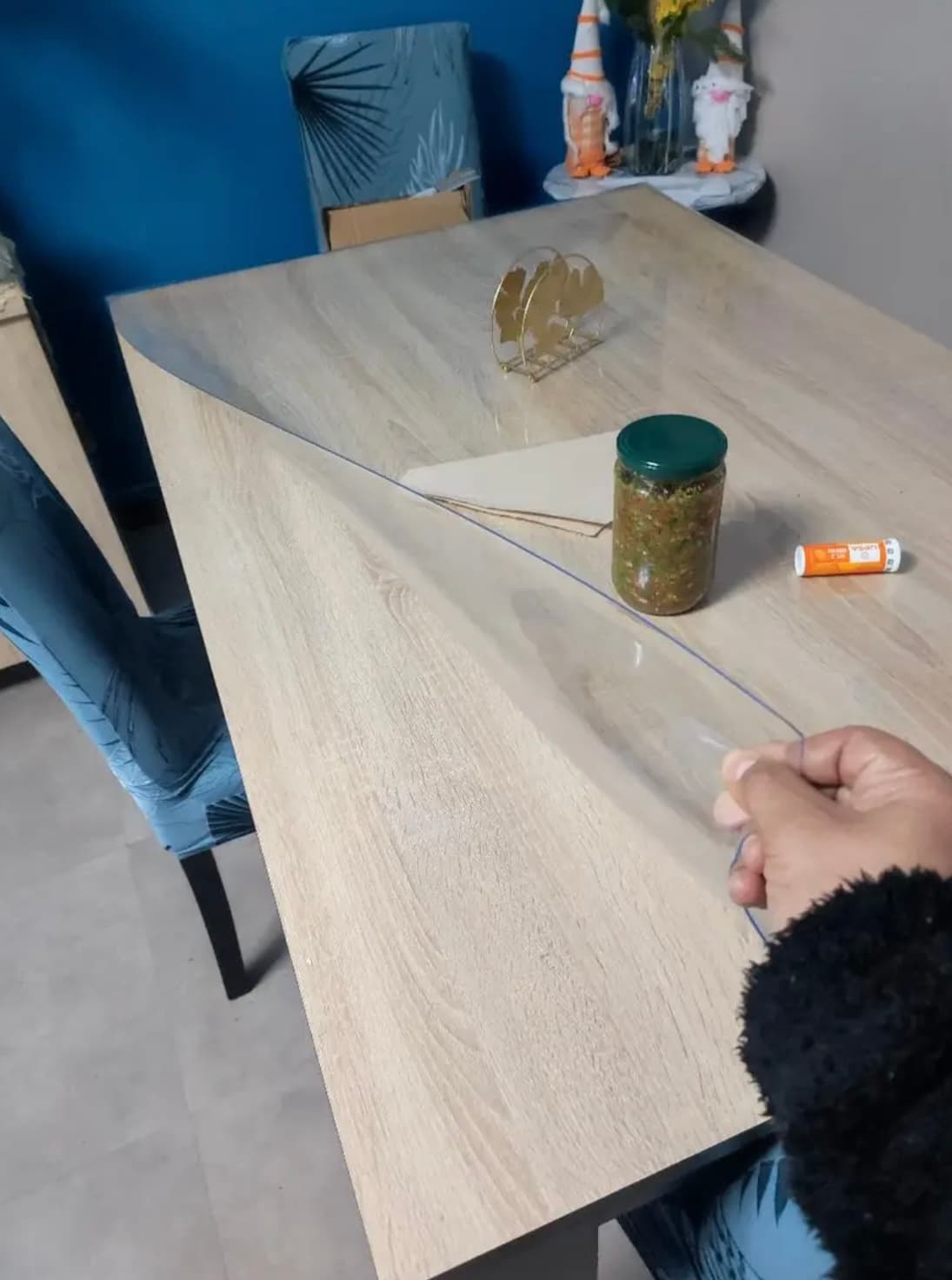 Eine Hand hebt die Ecke der transparenten Matte von einem Tisch aus heller Eiche; daneben steht ein Einmachglas