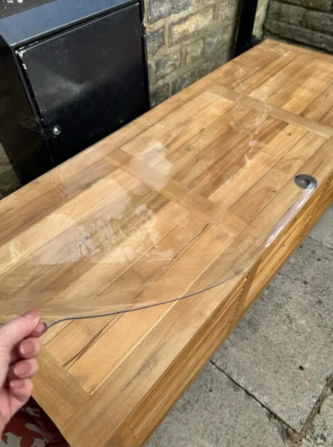 Gartenbank aus Teakleisten unter einer transparenten Matte; eine Hand hält deren Ecke