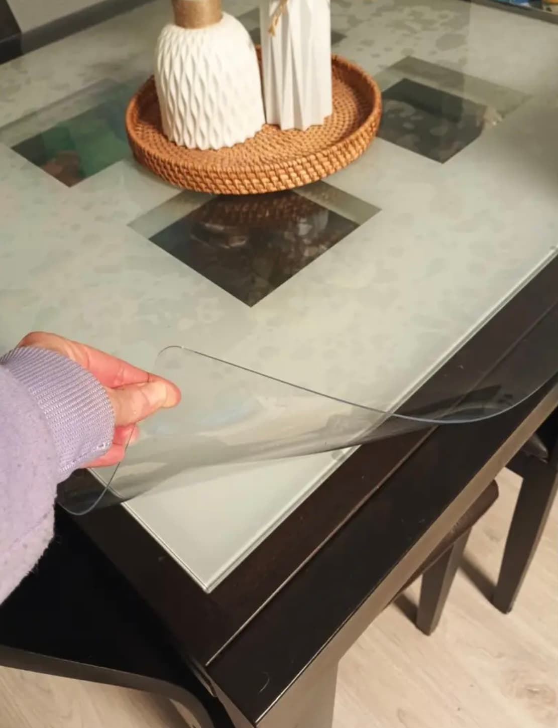 Eine Hand klappt die Ecke der Matte von einem dunklen Tisch mit matter Glasplatte und Rattantablett in der Mitte zurück