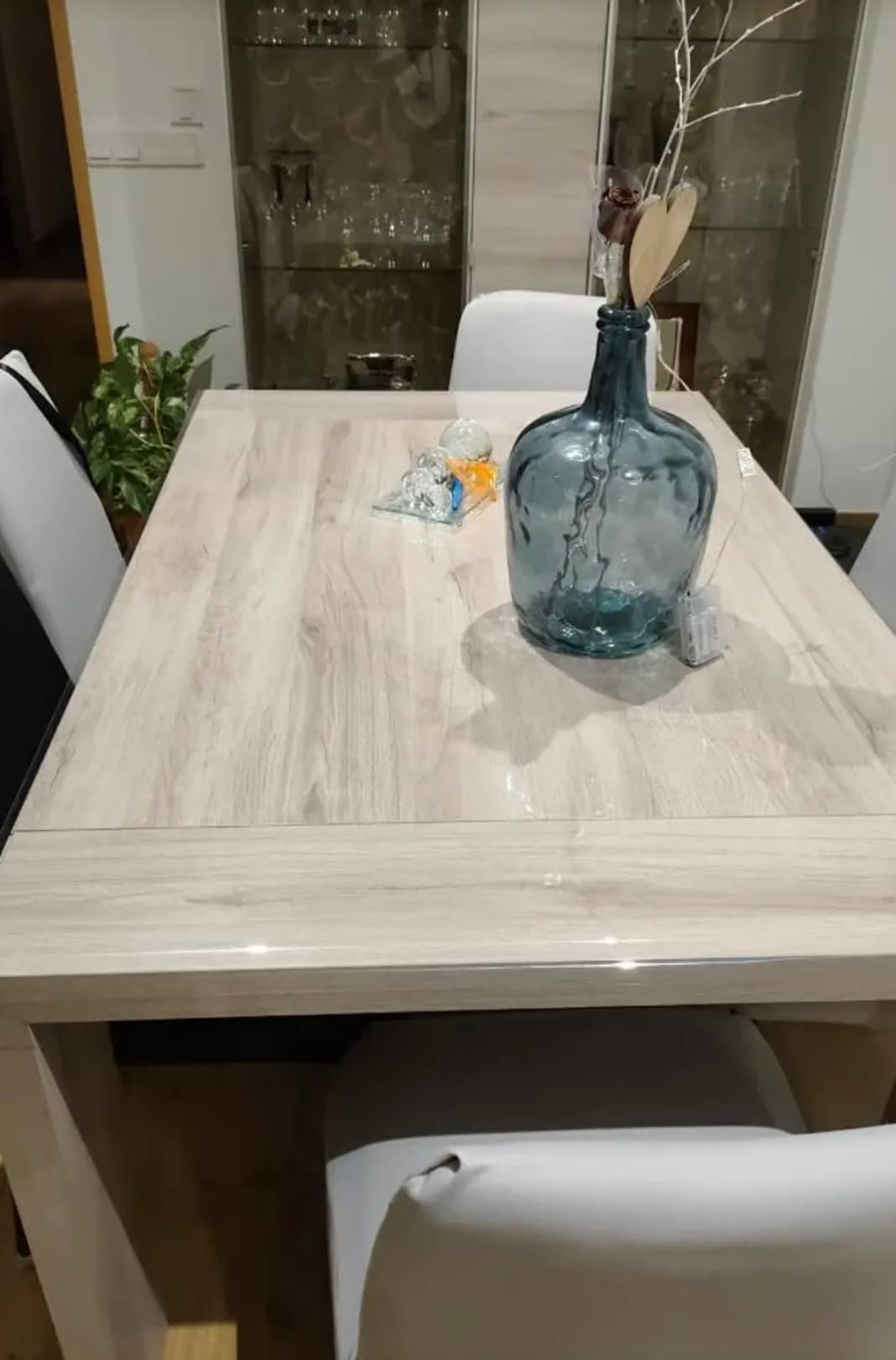 Ausziehtisch aus hellem Holz mit einer großen blauen Glasflasche; die Platte unter weichem Glas