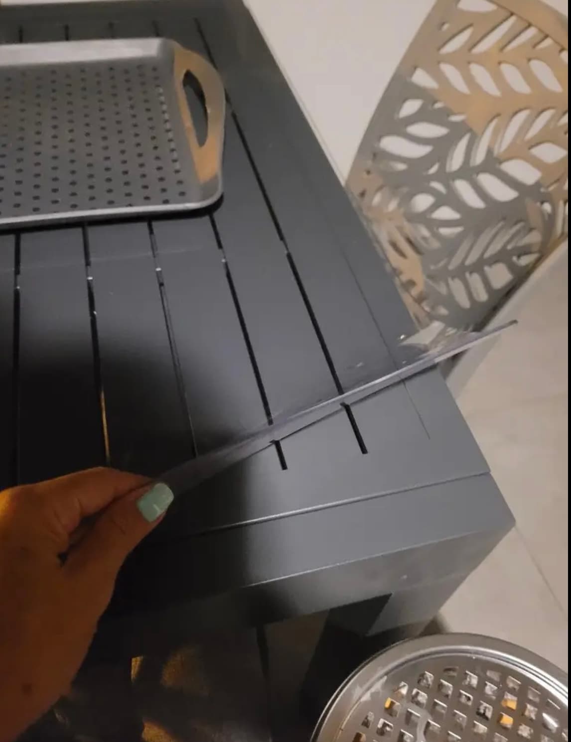 Eine Hand hebt die Ecke der Matte von einem anthrazitfarbenen Lattentisch, daneben stehen Metalltabletts