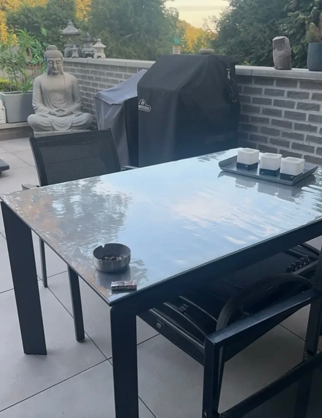 Terrasse mit Grill und Steinfigur; Tisch auf schwarzem Gestell, mit transparenter Matte bedeckt